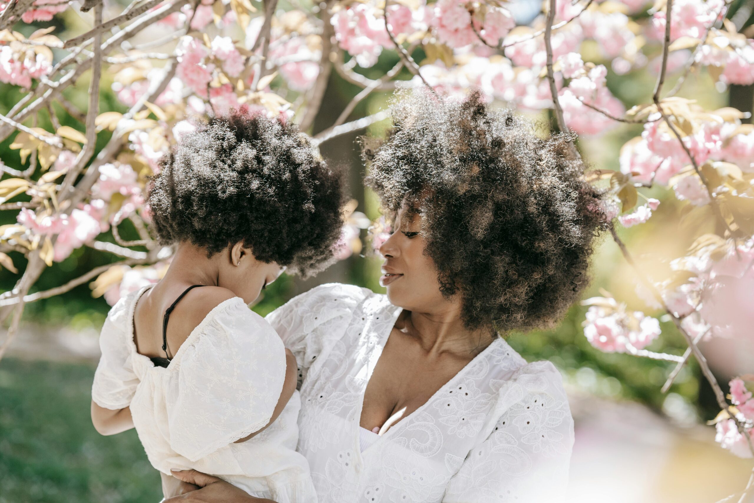 A young Black woman holds her daughter outside in front of a flowering tree.