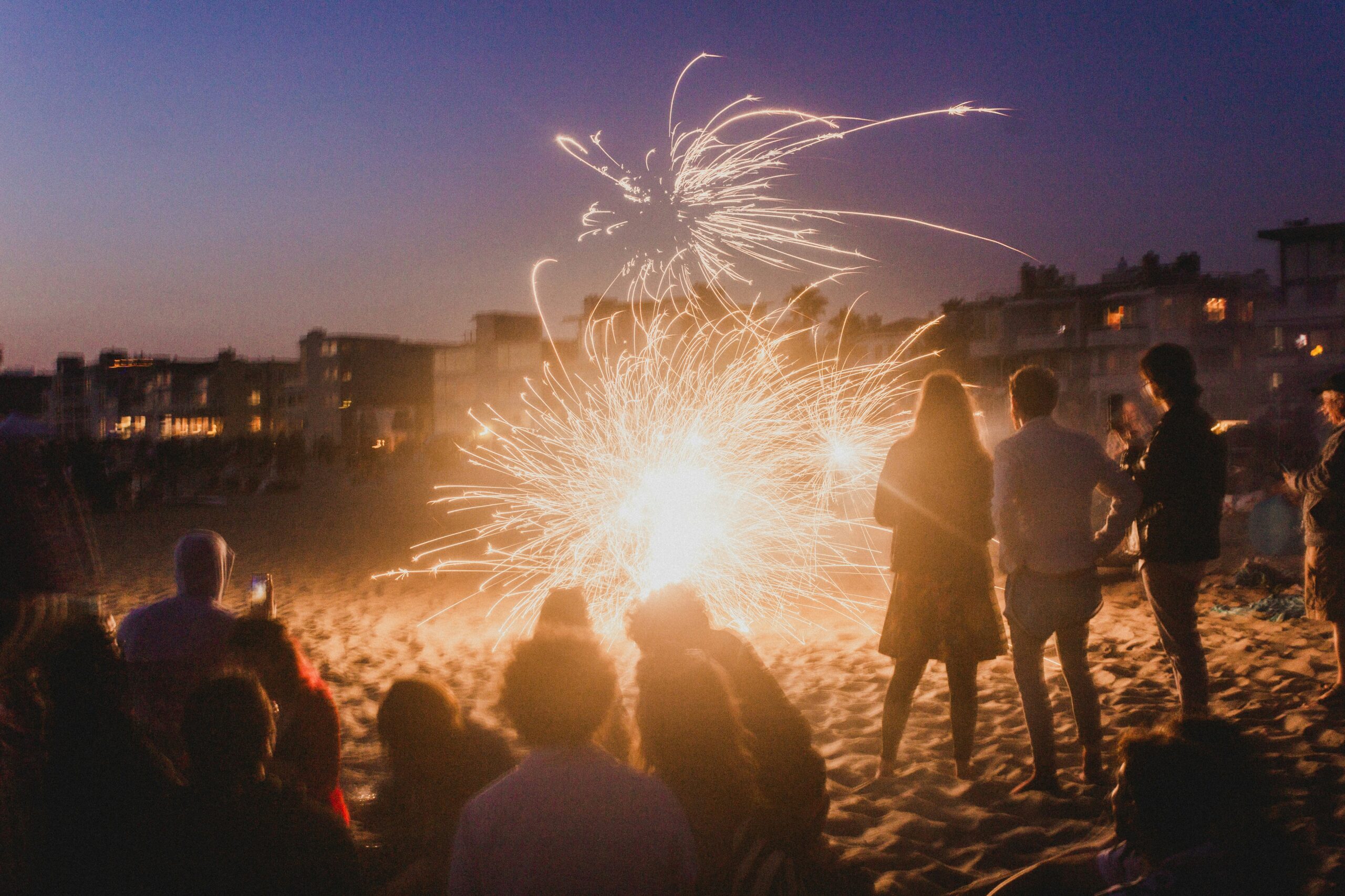 Group of people watching fireworks on the beach at dusk.
