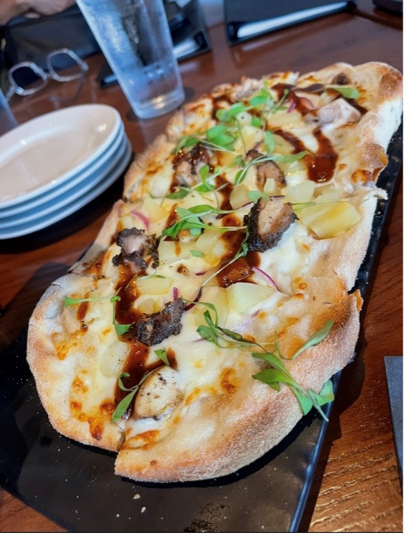 Closeup of a flatbread featuring meat, pineapple, cheese, and a drizzled sauce