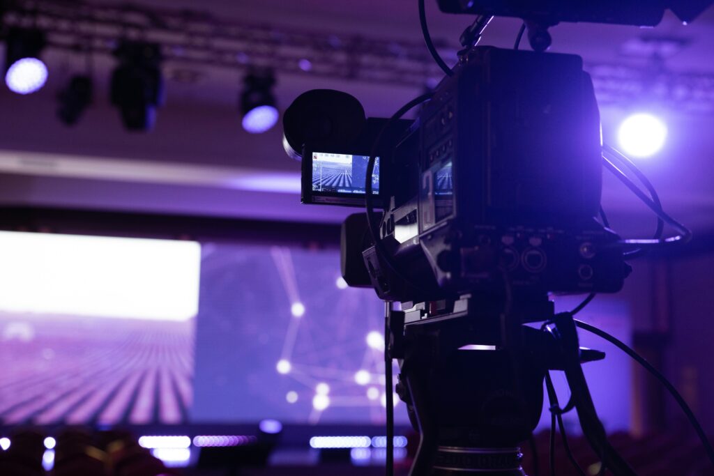 A video camera sits on a tripod, capturing a stage awash in purple lights.