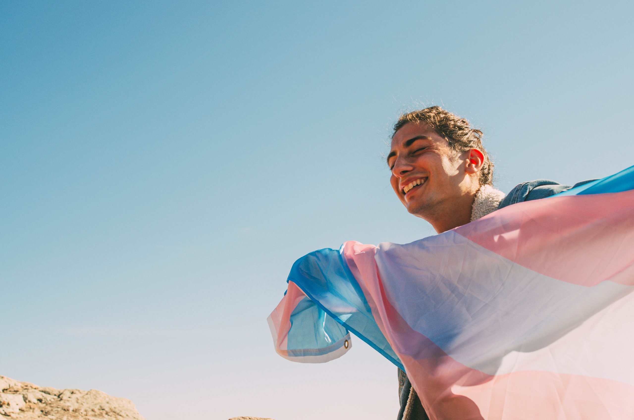 A trans-masculine young adult with curly brown hair smiling, holding the transgender pride flag.