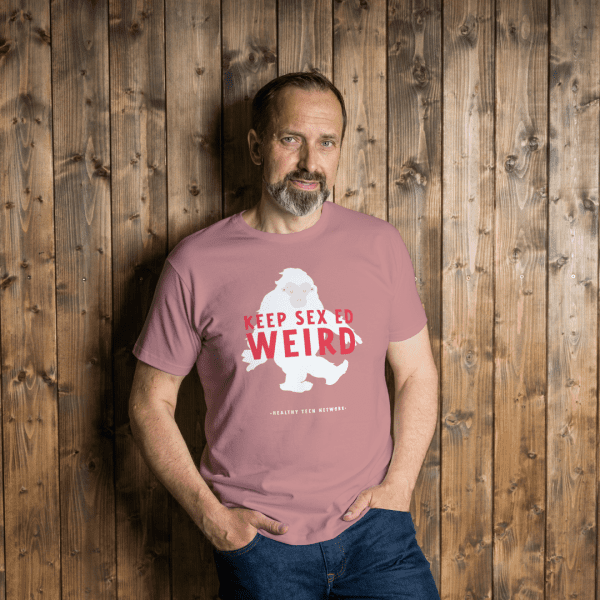 Man with hands in jean pockets wearing a dark pink t-shirt with a graphic of a sasquatch with the words "Keep Sex Ed Weird" in red written across