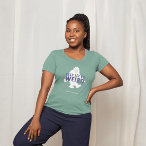woman wearing a ponytail wearing a sage green v-neck t-shirt with a graphic of a sasquatch with the words "Keep Sex Ed Weird" in purple written across