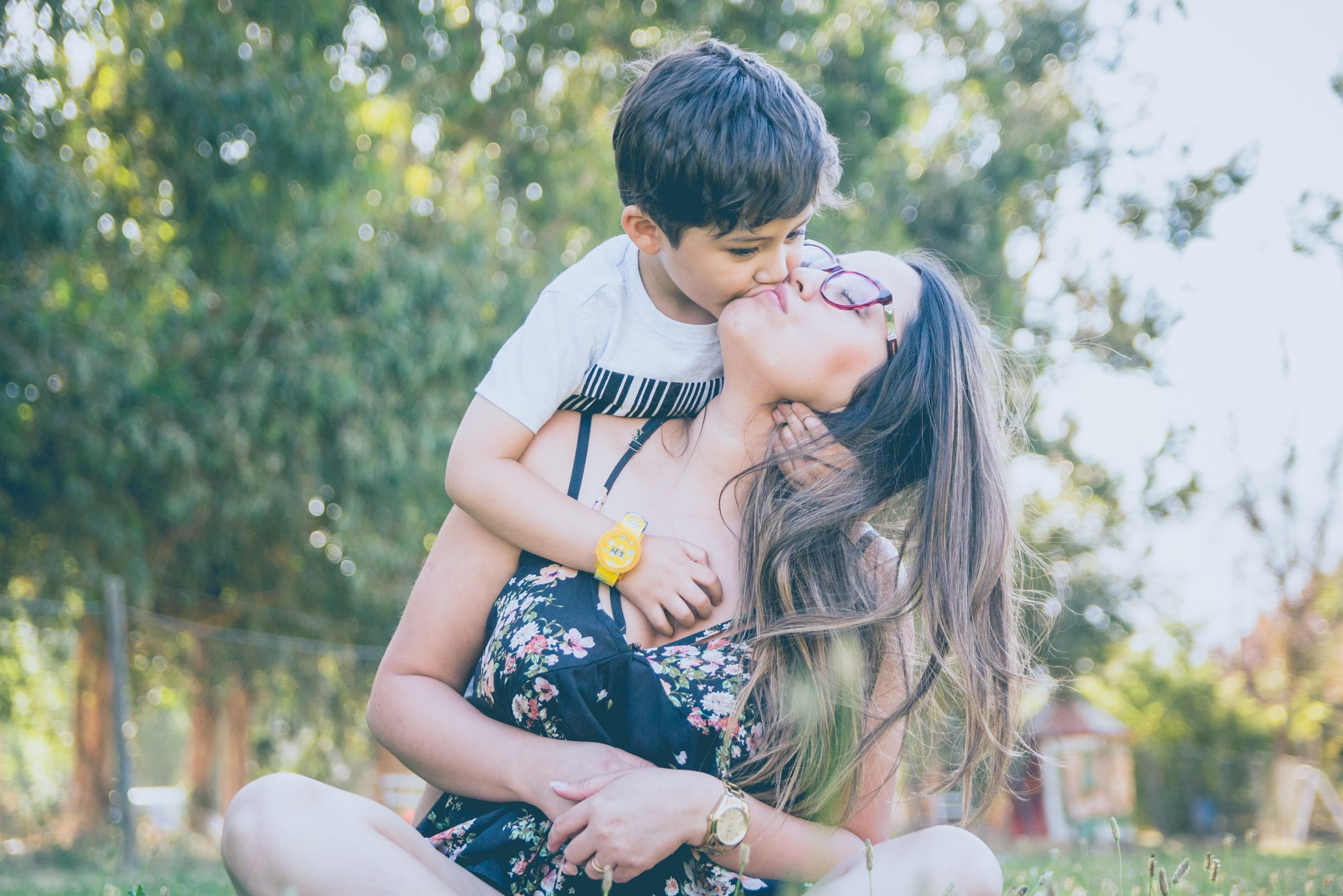 Mother sitting in a park with toddler son hanging on her back and giving her a kiss