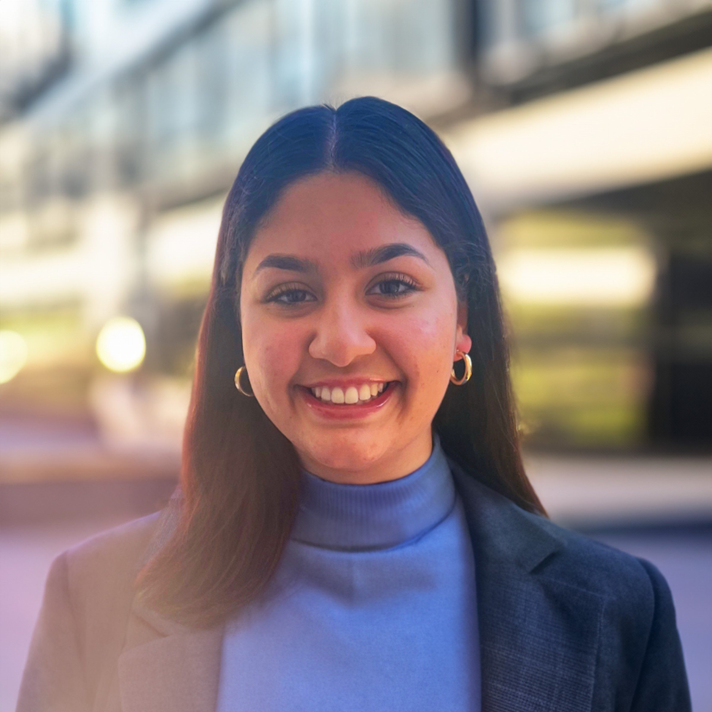 Photo of Asha Mohan from the shoulders up, smiling into the camera wearing a light blue turtleneck and navy blazer