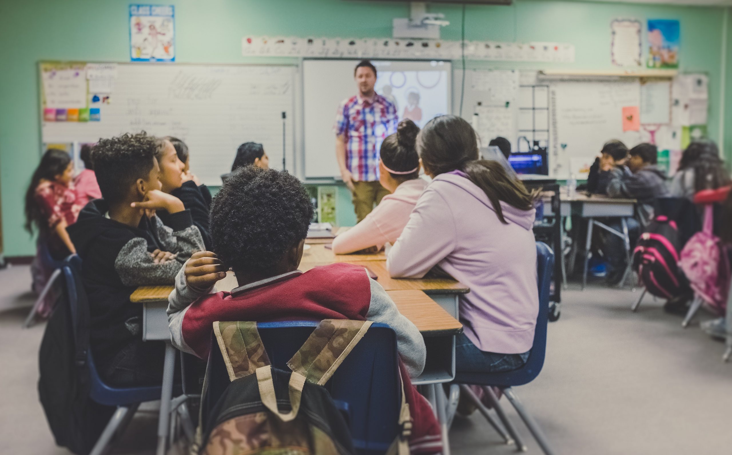 Teacher standing at front of classroom with students sat in groups infant of him